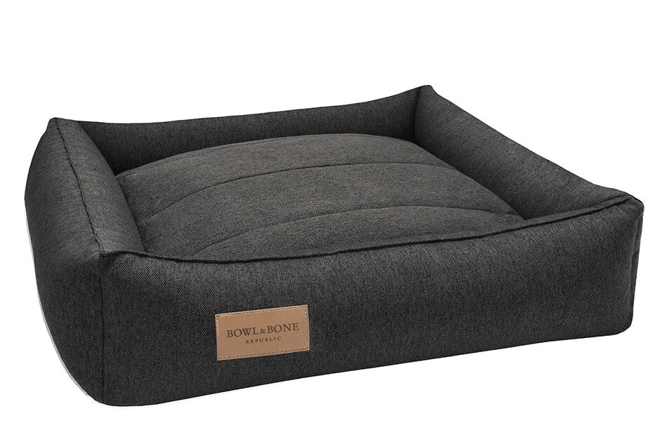 A grey dog bed with a brown leather tag by Bowlandbone, offering an URBAN graphite style.