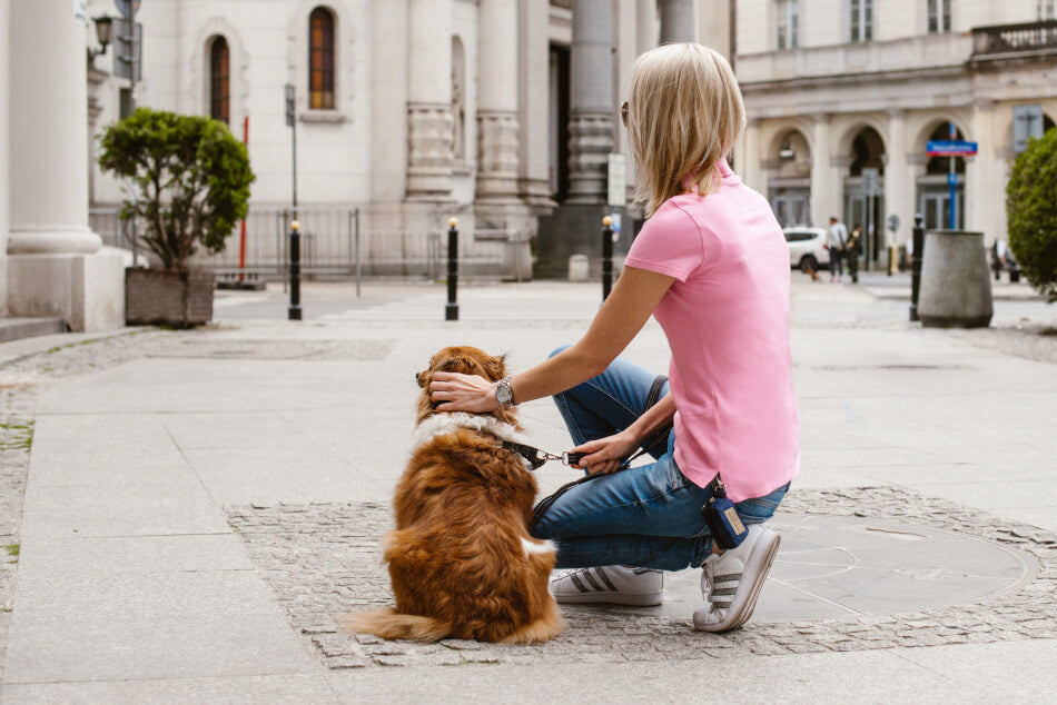 A woman kneeling down with her Bowlandbone dog waste bag holder MINI red in front of a Bowl&Bone Republic building.